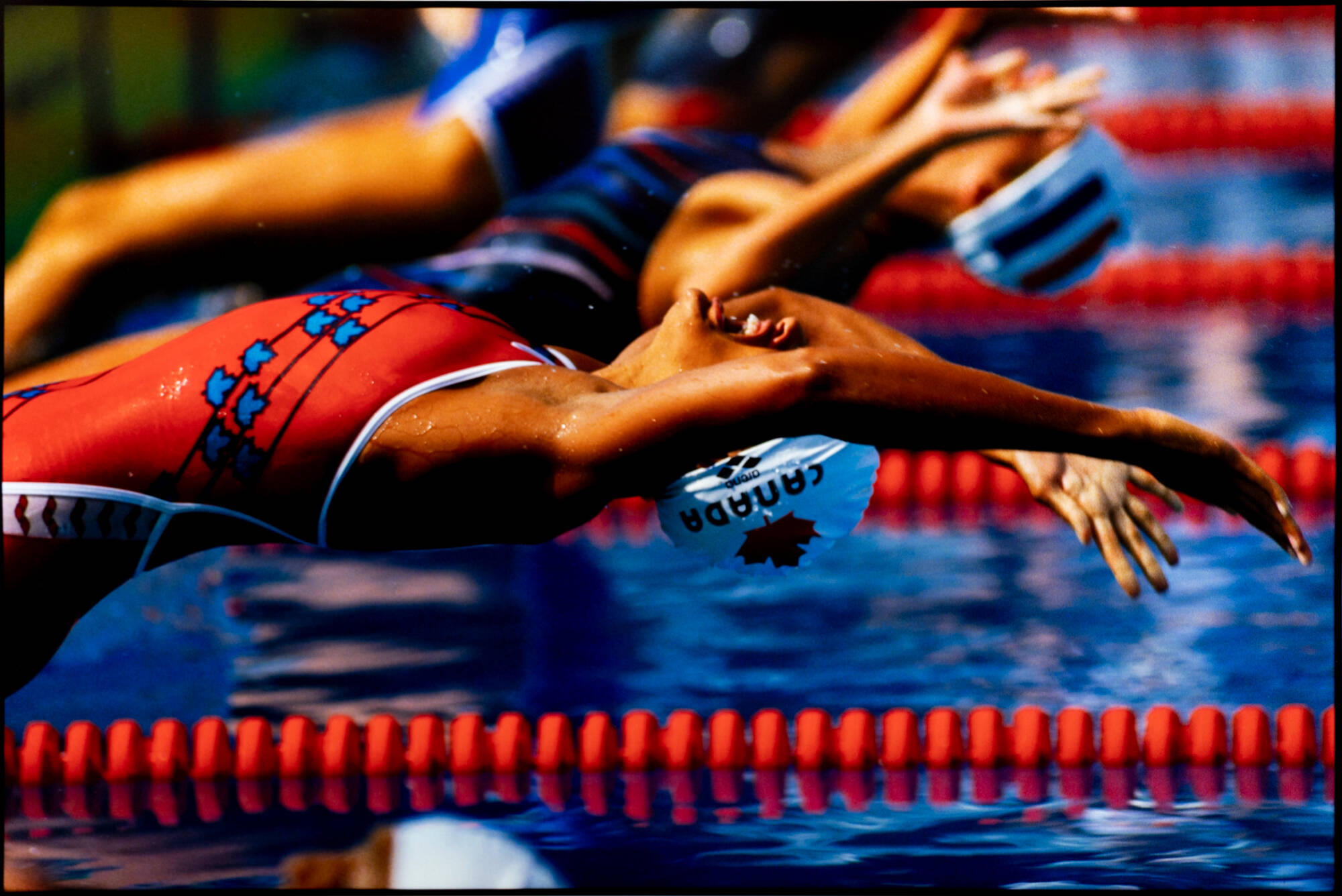 A horizontal color photograph of a swimmer. They are wearing a swim cap that reads "Canada" and a red swimsuit.
